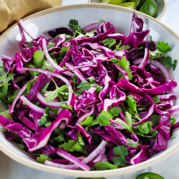 A bowl filled with a vibrant salad of shredded purple cabbage, sliced red onion, and fresh cilantro leaves, with lime wedges visible in the background.