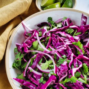 A bowl of fresh purple cabbage salad mixed with cilantro, served next to lime wedges and a mustard-colored cloth napkin.