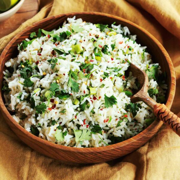 A wooden bowl filled with cilantro lime rice mixed with chopped green onions, garnished with red pepper flakes, sits on a brown cloth with a wooden serving spoon resting inside.