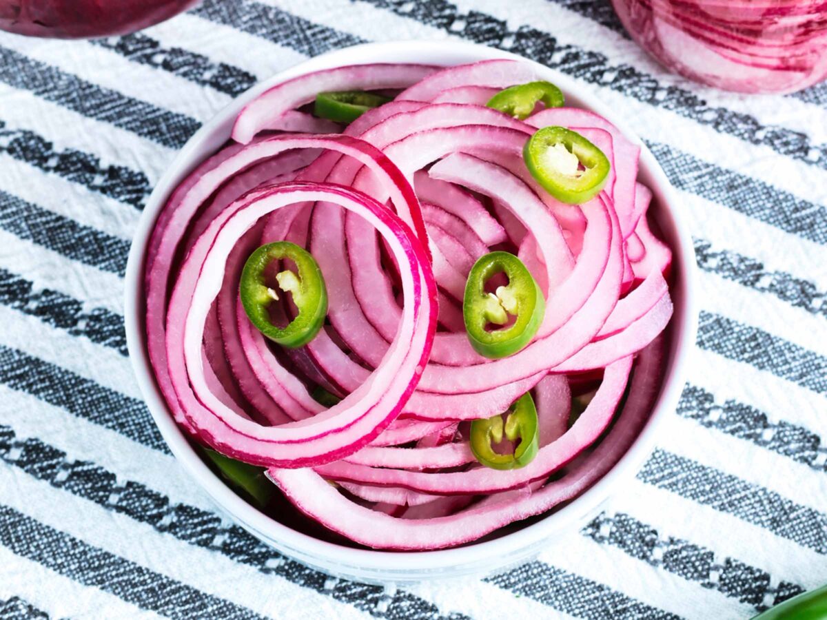 A white bowl filled with quick pickled red onions and green jalapeño peppers sits on a black and white striped cloth.