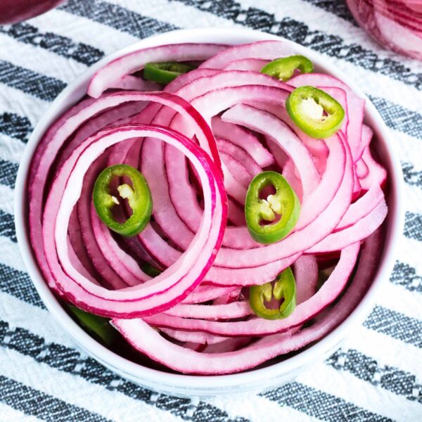 A white bowl filled with quick pickled red onions and green jalapeño peppers sits on a black and white striped cloth.
