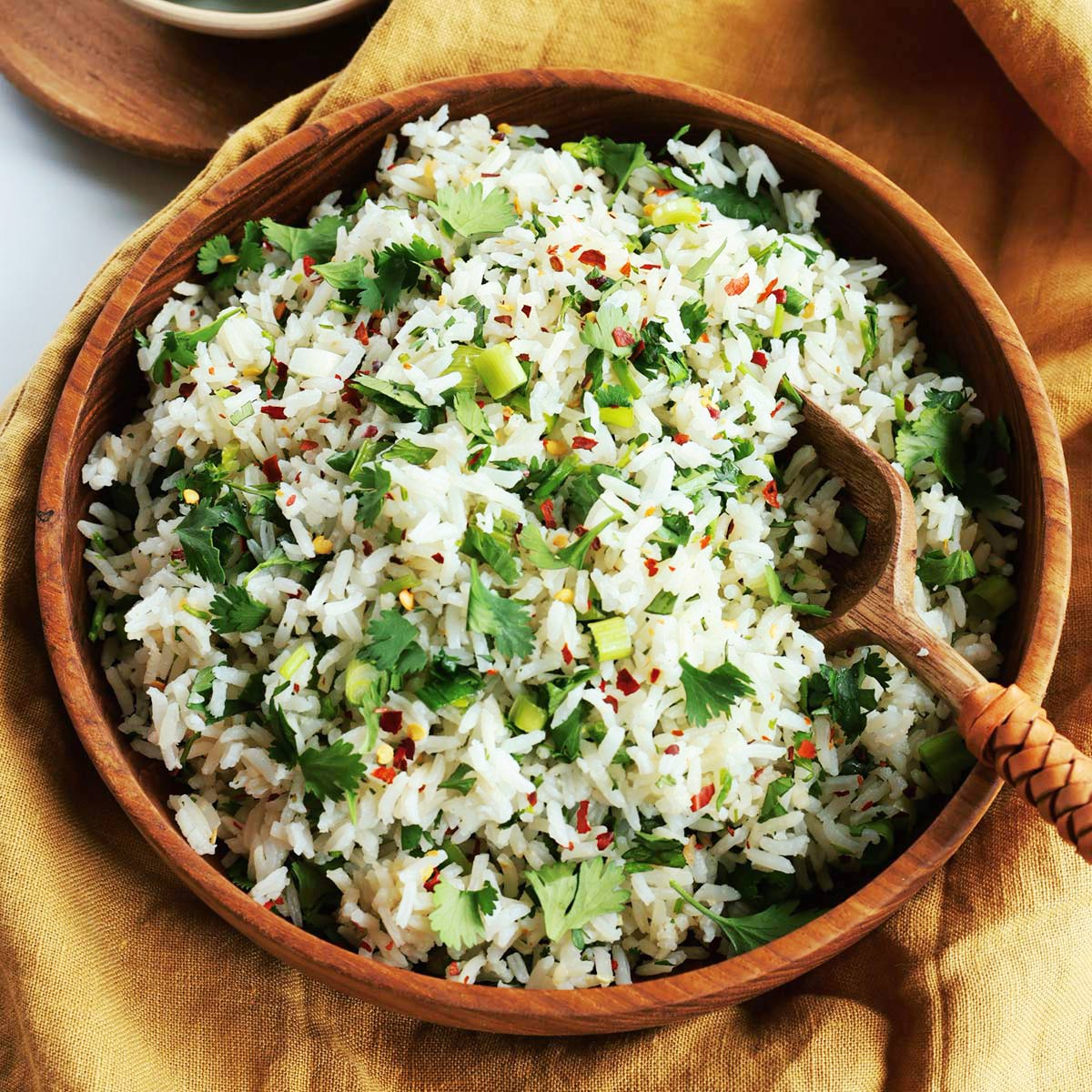 A wooden bowl filled with cilantro lime rice, mingled with chopped green onions and red pepper flakes, rests atop a mustard-colored cloth. A wooden serving spoon is nestled inside.