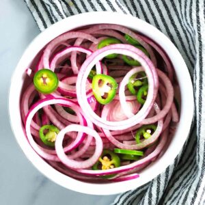 A white bowl filled with quick pickled red onions and green jalapeño peppers, placed on a striped cloth. The vegetables are arranged in overlapping rings and slices.