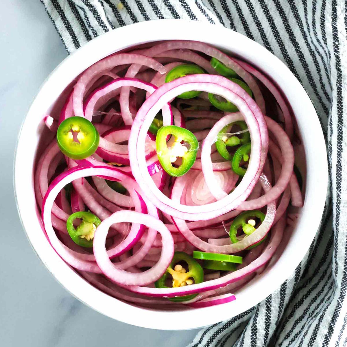 A white bowl filled with quick pickled red onions and green jalapeño peppers, placed on a striped cloth. The vegetables are arranged in overlapping rings and slices.