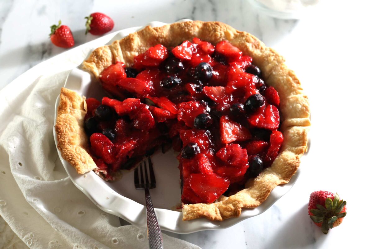 A fruit pie with a golden crust, filled with juicy strawberries and blueberries, sits on a white plate. A slice has been removed, revealing the vibrant filling. Whole strawberries are scattered nearby on the marble surface for an extra fruity touch.