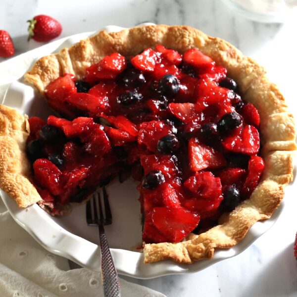 A fruit pie with a golden crust, filled with juicy strawberries and blueberries, sits on a white plate. A slice has been removed, revealing the vibrant filling. Whole strawberries are scattered nearby on the marble surface for an extra fruity touch.