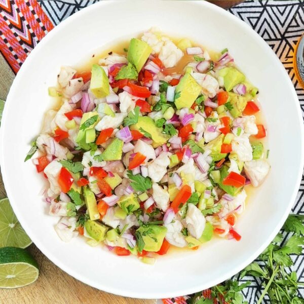 A bowl of ceviche with fish, avocado, red onion, tomato, cilantro, and lime, surrounded by fresh lime halves, herbs, and patterned textiles on a wooden surface.